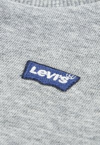 Γκρι πλεκτό ύφασμα με κεντημένη μπλε ετικέτα λογότυπου Levi's με λευκά γράμματα, τοποθετημένη στο κέντρο. Απαλή υφή και casual στυλ.