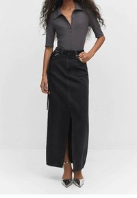 Gonna maxi in denim nero con spacco frontale, abbinata a una polo a coste grigia e tacchi a punta argentati. Texture liscia e design aderente.