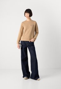 Donna in jeans a gamba larga scuri, felpa beige con logo, tiene in mano una borsa argentata su uno sfondo bianco uniforme.