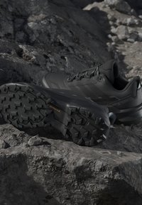 Scarpe da trekking nere con suole profondamente scolpite posizionate su terreno roccioso, progettate per un uso esterno impegnativo.