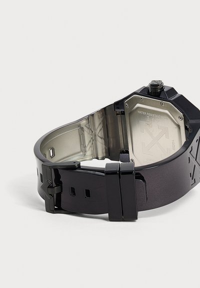 Montre-bracelet noire avec un bracelet partiellement ouvert révélant le couvercle arrière en métal marqué comme résistant à l'eau et fixé avec des vis.