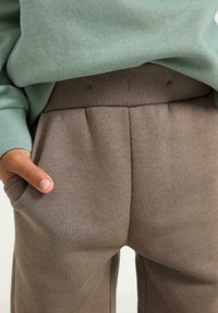 Grüner Pullover mit gerippten Bündchen, kombiniert mit braunen Hosen, die seitliche Taschen und subtile Falten an der Taille aufweisen. Weiche Stofftextur.
