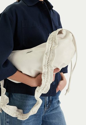 Osoba w ciemnej kurtce i dżinsach trzyma miękką, kremową torebkę z marszczonego materiału z długim paskiem i zamknięciem na ściągacz.