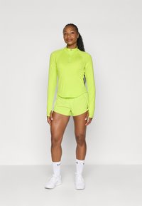 Limegrünes sportliches Langarmoberteil mit halbem Reißverschluss, kombiniert mit passenden Shorts. Enthält das Nike-Logo, hat eine glatte Textur und eine figurbetonte Passform.