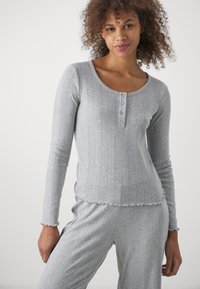 Haut gris à manches longues à côtes avec un décolleté en cœur et un détail de quatre boutons, présentant des bords effilochés et un tissu doux et texturé.