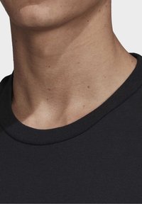 Černé bavlněné tričko s kulatým výstřihem, viditelným šitím a hladkou strukturou; detailní pohled na oblast krku a ramene.