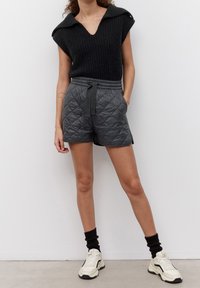 Haut sans manches en tricot noir avec un col en V, associé à un short tombant en gris matelassé avec une taille élastique et des poches latérales. Sneakers blanches et chaussettes noires.
