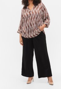 Donna che indossa pantaloni neri larghi, tacchi alti beige e una camicia a maniche lunghe a motivi marroni e bianchi con scollatura a V, con una mano in tasca.
