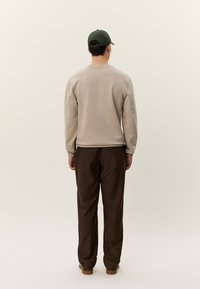 Hombre con sudadera beige, pantalones marrón oscuro y gorra de béisbol verde, de pie de espaldas contra un fondo claro y liso.