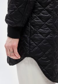 Αριστερό χέρι με ασημένιο δαχτυλίδι με χάντρες κρεμασμένο δίπλα σε μαύρο πουπουλένιο μπουφάν με ριμ π μανσέτα και ανοιχτόχρωμο παντελόνι.