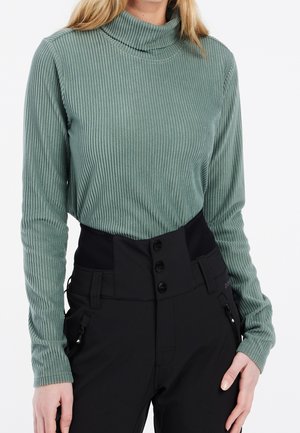 Kobieta nosząca prążkowany zielony sweter z golfem wpuszczony w czarne spodnie z wysokim stanem, z zapięciem na guziki i kieszeniami na suwak.