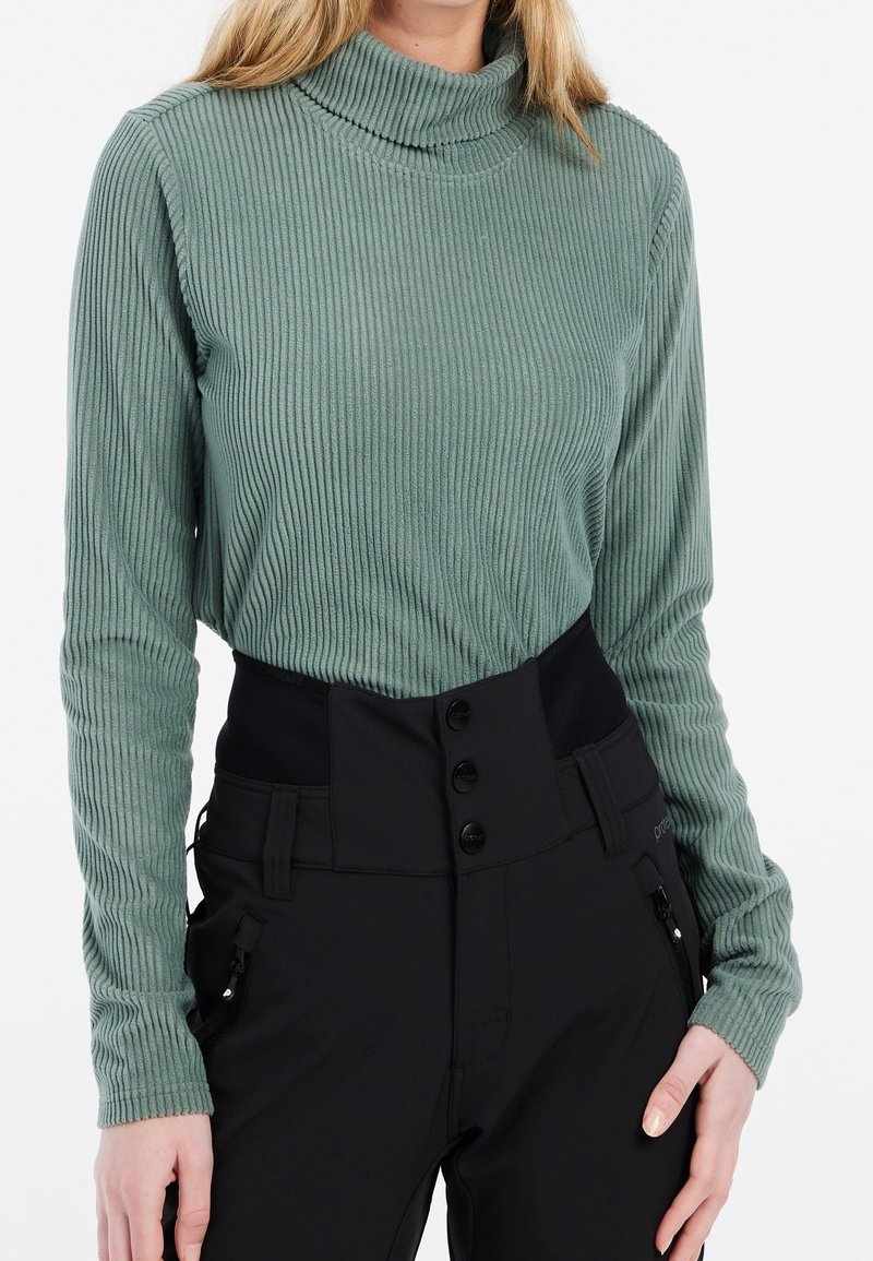 Femme portant un pull à col roulé vert côtelé rentré dans un pantalon noir taille haute avec fermeture par boutons et poches zippées.