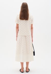 White embroidered top with short sleeves, paired with a matching skirt that has a smooth hem. Model wears black slide shoes and holds a small bag.