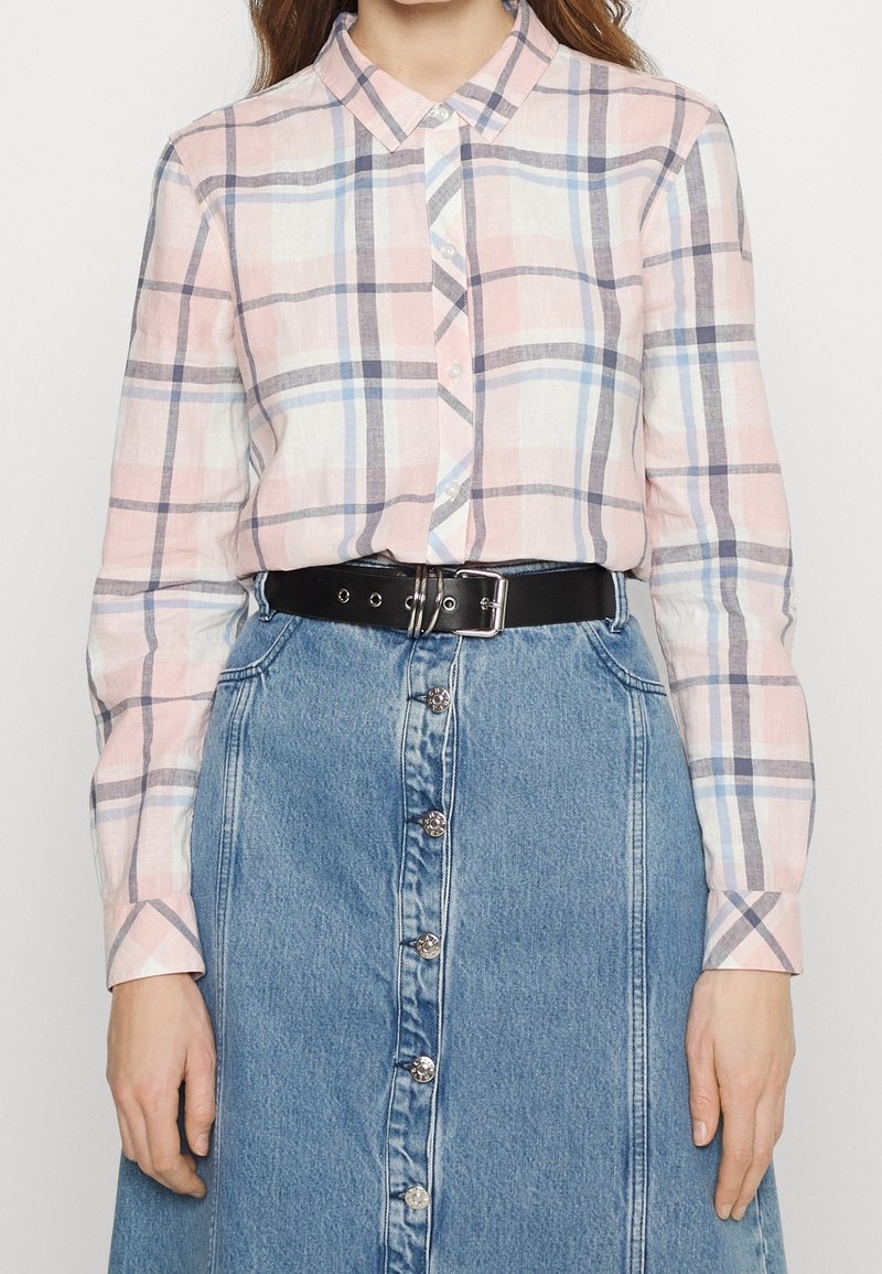 Femme portant une chemise à carreaux rose et bleue boutonnée, rentrée dans une jupe en jean bleue à boutons sur le devant avec une ceinture noire.