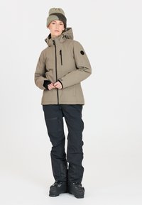 Ski-jacka i ljus khaki med dragkedja fram och justerbar huva. Svarta skidbyxor med dragkedjor, i kombination med svarta skidkängor och en grön stickad mössa.