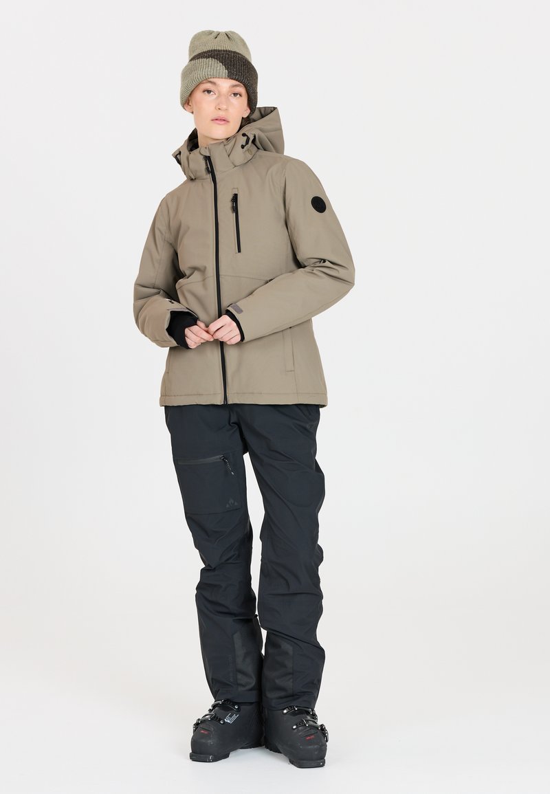 Ski-jacka i ljus khaki med dragkedja fram och justerbar huva. Svarta skidbyxor med dragkedjor, i kombination med svarta skidkängor och en grön stickad mössa.