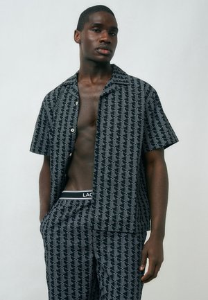 Jeune homme portant une chemise à manches courtes et un pantalon assortis à motifs noirs et blancs, les mains dans les poches, sur un fond uni.