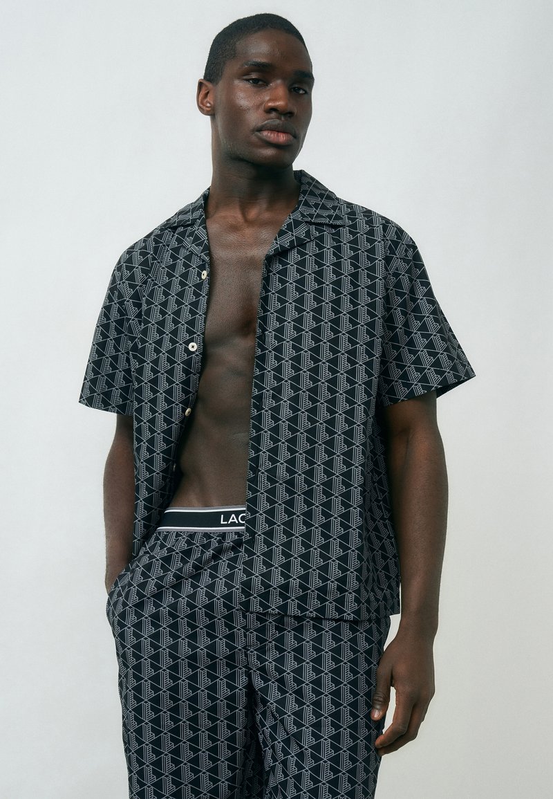 Νεαρός άνδρας που φοράει μαύρο και λευκό πουκάμισο με κοντά μανίκια και παντελόνι με το ίδιο μοτίβο, με τα χέρια στις τσέπες, μπροστά σε απλό φόντο.