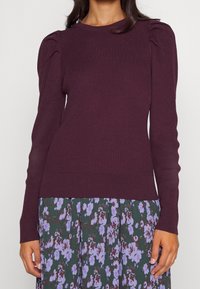 Burgundy ribbad stickad tröja med lång ärm och puffiga axlar, parat med en blomstermönstrad kjol i lila och gröna nyanser.