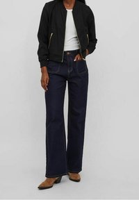 Veste bomber noire avec des zips dorés, haut ajusté blanc et jeans taille large bleu foncé. Des bottines à talons marron complètent l'ensemble.