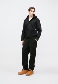 Jachetă ușoară neagră cu glugă și două buzunare frontale, combinată cu pantaloni din catifea cu reiat verde închis și cizme căptușite de culoare cărămizie, cu șireturi.