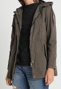 Parka verde oliva con cierre de cremallera y botones a presión, capucha ajustable y dos bolsillos frontales. Tela suave y texturizada con un ajuste casual.