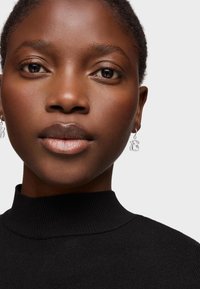 Suéter negro de cuello alto con detalles acanalados, acompañado de pendientes plateados con un diseño geométrico, resaltando la piel suave y un tono de labios natural.