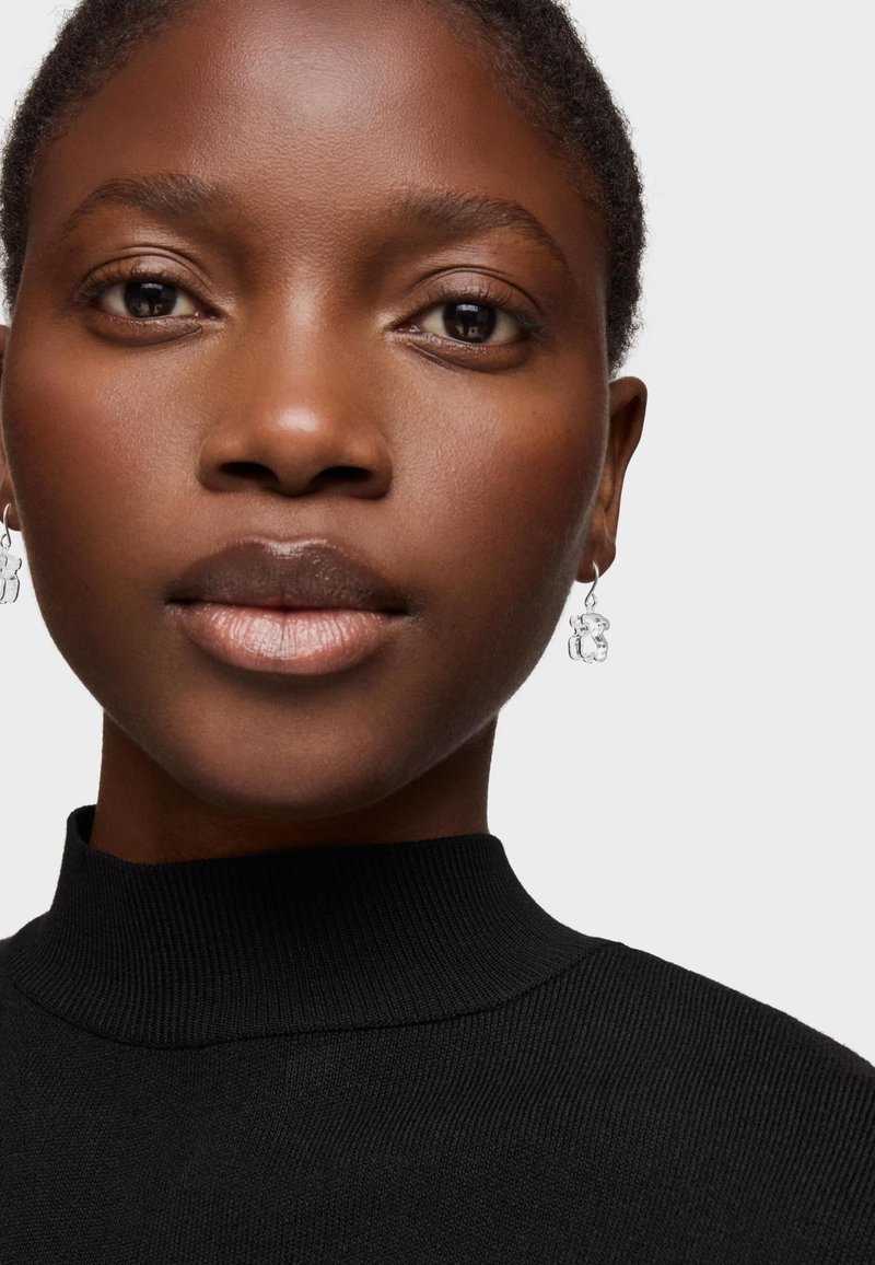 Suéter negro de cuello alto con detalles acanalados, acompañado de pendientes plateados con un diseño geométrico, resaltando la piel suave y un tono de labios natural.