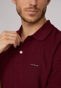 Polo bordeaux avec un col classique, un bouton simple et trois motifs d'animaux gris sur la poitrine gauche. Texture en coton doux.