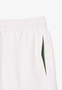 Pantaloni corti rosa chiaro con cintura elastica, dotati di tasche laterali foderate in verde. Texture liscia e design minimalista.