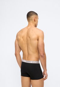 Μυώδης άνδρας με κοντά μαλλιά που φοράει μαύρα μποξεράκια Tommy Hilfiger, στέκεται πλάτη με φόντο ένα απλό ανοιχτόχρωμο φόντο.