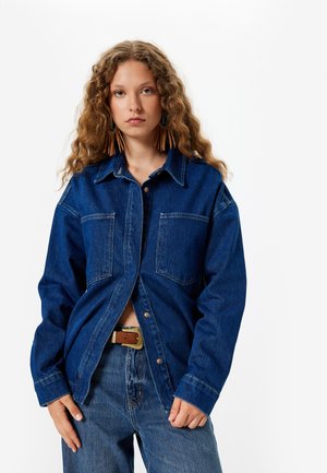 Das Jeanshemd hat eine dunkelblaue Farbe, eine durchgehende Knopfleiste, zwei Brusttaschen und einen spitzen Kragen, kombiniert mit hellblauen Jeans und einem braunen Gürtel.