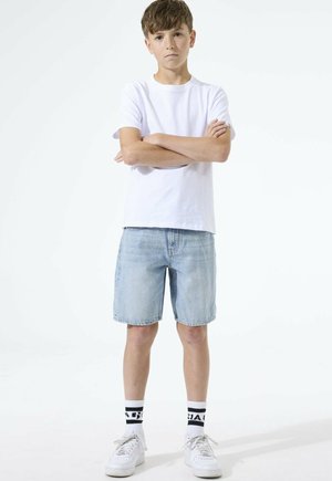 Ragazzo con capelli castani corti in piedi a braccia conserte, indossa una maglietta bianca, pantaloncini di denim chiaro, calzini bianchi con scritte nere e scarpe da ginnastica bianche.