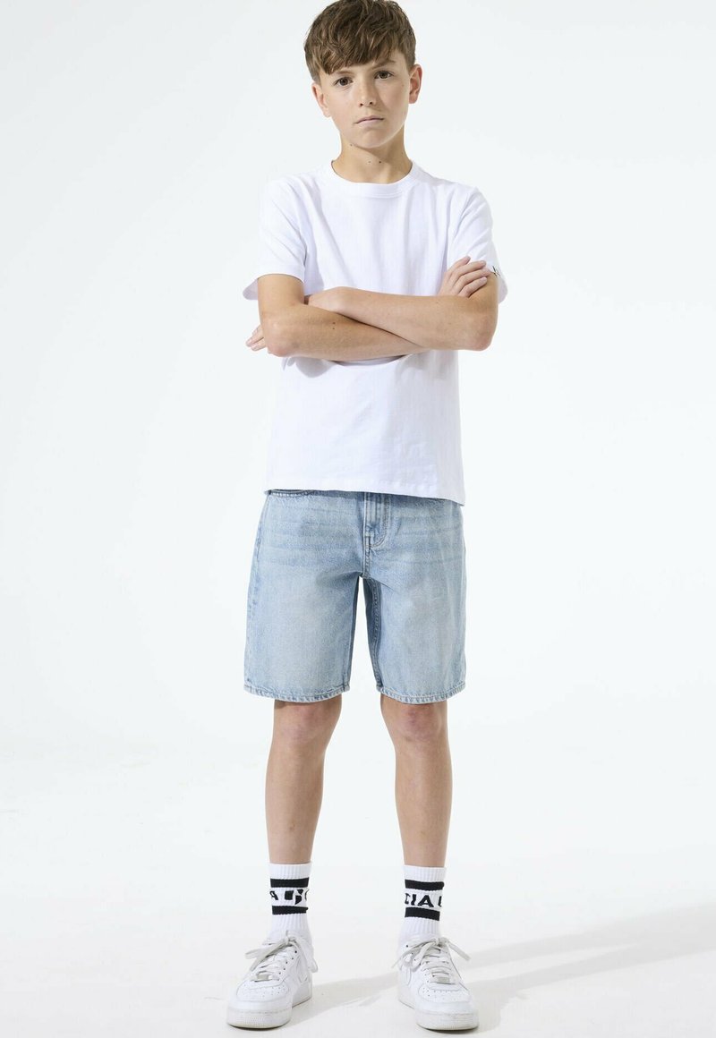 Niño con cabello corto y castaño, de pie con los brazos cruzados, vistiendo una camiseta blanca, pantalones cortos de mezclilla claros, calcetines blancos con texto negro y zapatillas blancas.