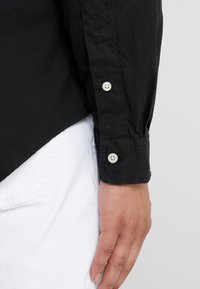 Černá košile s dlouhým rukávem a manžetami na knoflíky, vyrobená z lehké látky. Má mírně zaoblený lem a jemnou texturu, kombinovaná s bílými kalhotami.