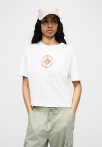 Νεαρός με μακριά σκούρα μαλλιά φοράει λευκό μπλουζάκι Columbia Sportswear, μπεζ καπέλο και ανοιχτό πράσινο παντελόνι με κορδόνι, με τα χέρια στις τσέπες.