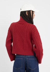 Czerwony sweter robiony na drutach z wzorami w warkocze, z wysokim kołnierzem i skróconym fasonem. Posiada ściągacz na dolnej krawędzi i rękawach. Zestawiony z niebieskimi dżinsami.
