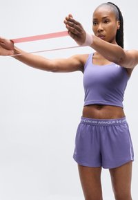 Αθλητική γυναίκα με μωβ ρούχα γυμναστικής τεντώνει μια ροζ λάστιχο αντίστασης με συγκεντρωμένη έκφραση σε λευκό φόντο.