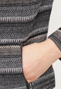 Šedá tkanina s vzorem a texturovaným designem v zigzag, zobrazující detail na ruku v bočním kapse. Zipový detail na spodním lemu.