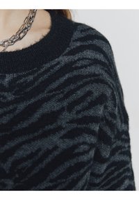 Pull en tricot noir et gris avec un motif de rayures de zèbre, col rond et texture douce. Collier en chaîne argenté visible.