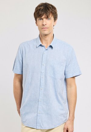 Homme debout, faisant face à l'avant, portant une chemise à manches courtes boutonnée bleu clair et un pantalon beige, sur un fond uni.