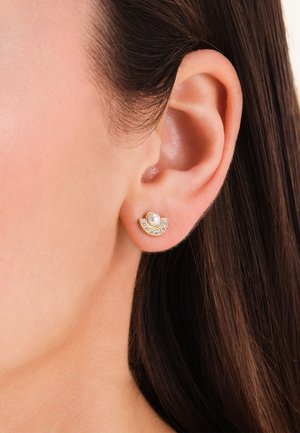 Close-up of a woman's ear wearing a gold stud earring with a central pearl and a curved row of small clear stones.