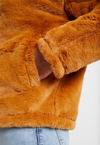Umělá kožešinová oranžová bunda s měkkou strukturou, obsahující boční kapsu a manžety na rukávech. Tkanina je sametová a silná, poskytující teplo.