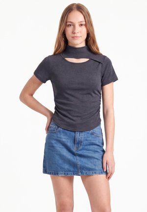 Junge Frau mit langen Haaren, die ein dunkelgraues kurzärmeliges Top mit Ausschnitt und einen blauen Jeansrock trägt, steht mit einer Hand in der Hüfte.