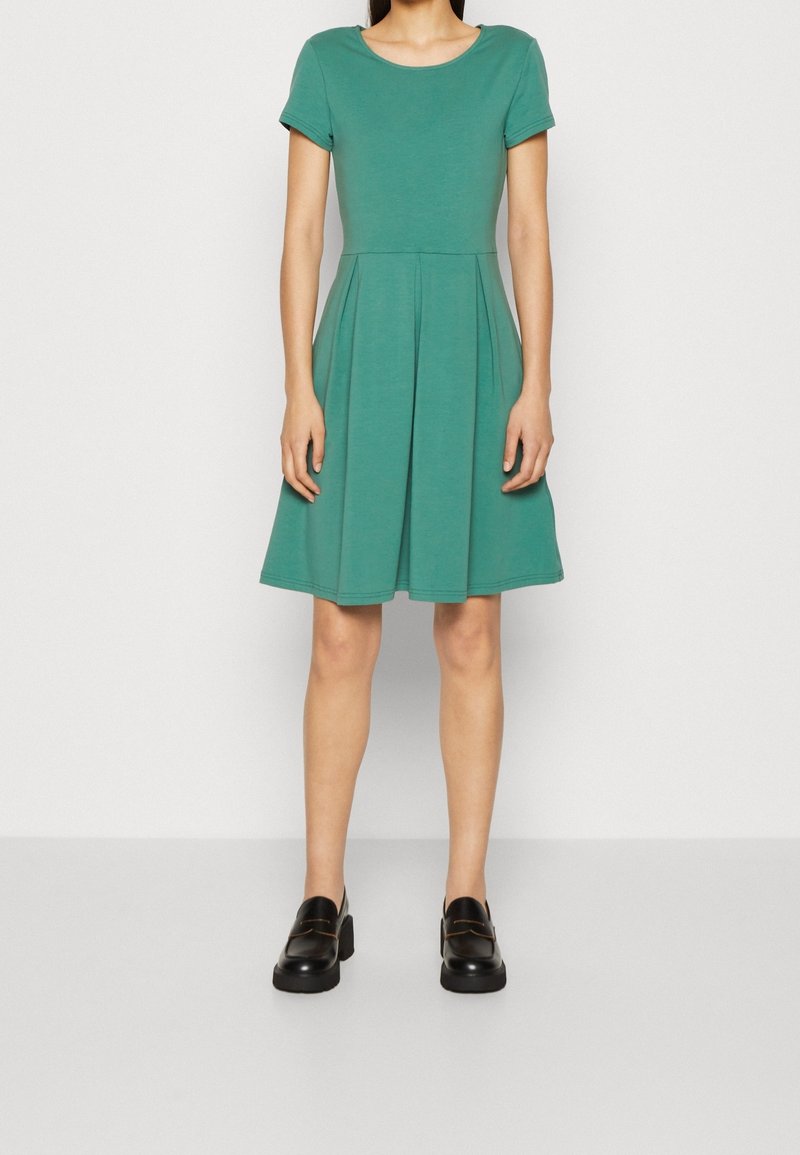 Zielona sukienka z krótkim rękawem, dopasowanym gorsetem i rozszerzaną spódnicą. Wykonana z miękkiego materiału, z plisami i okrągłym dekoltem. Czarne buty na platformie.