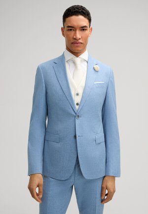 Uomo che indossa un abito azzurro chiaro con gilet bianco, cravatta e fiore all'occhiello, in piedi su uno sfondo grigio neutro.