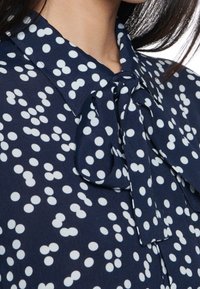 Blusa navy con pois bianchi, dotata di colletto e un dettaglio di fiocco annodato al collo. Tessuto morbido con una leggera trama.
