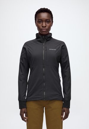 Une femme porte une veste Patagonia noire zippée à col montant et un pantalon marron, sur un fond blanc uni.