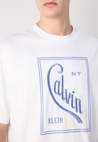 Balta kokvilnas T-kreklu ar zilu izšūtu "Calvin Klein" logotipu un taisnstūra kontūru, ar apaļu izgriezumu un īsām piedurknēm.