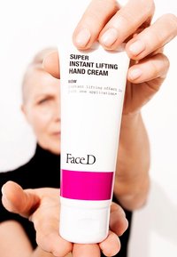 Tubo bianco di crema per le mani con una striscia rosa. Testo: "Crema per le mani Super Instant Lifting" e "FaceD." Tenuto in una mano con le dita visibili.